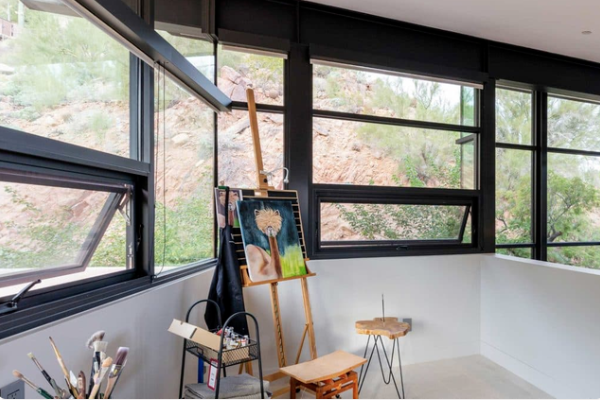 Easel and paints inside room with windows sporting thick black aluminum frames outlooking a beautiful garden